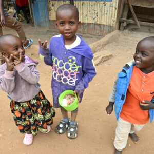 Three young children smiling, one holding a green bowl of food