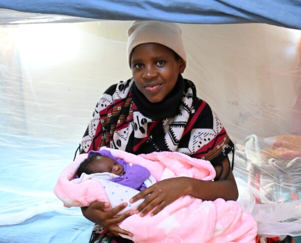 A young mother holding her newborn wrapped in a pink blanket beside a mosquito net
