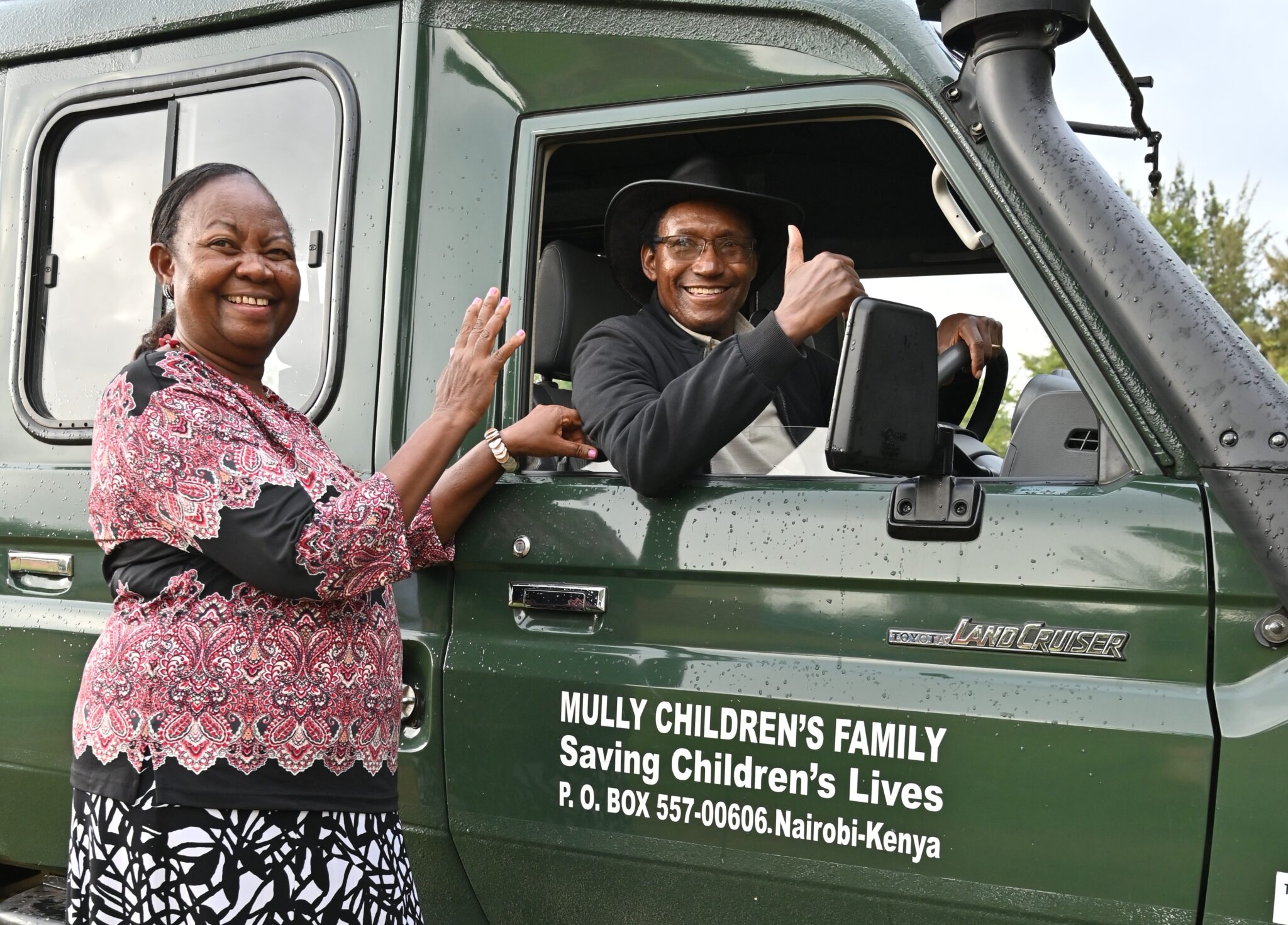 A smiling woman beside an MCF Land Cruiser while the driver gives a thumbs-up